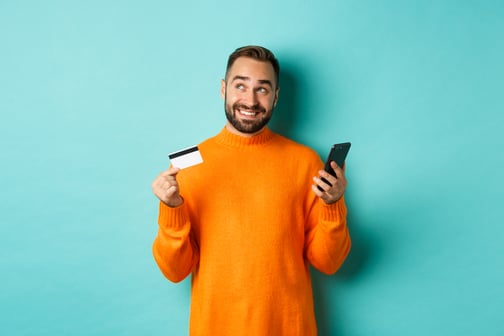 online-shopping-handsome-man-thinking-holding-smartphone-with-credit-card-paying-in-internet-store-standing-over-light-turquoise-wall